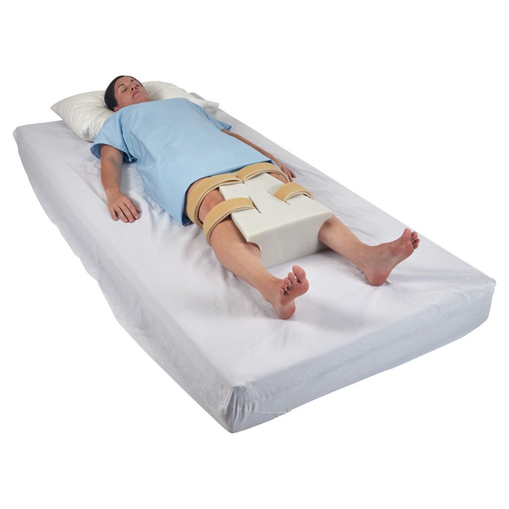 Person lying on back with a white foam abduction pillow secured between their thighs after hip surgery